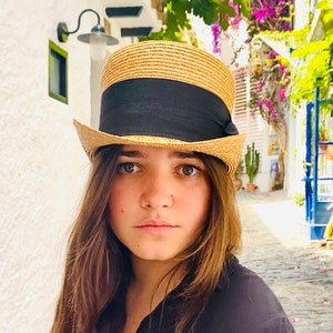 Peut inclure: Une femme aux longs cheveux bruns porte un chapeau de paille avec un bandeau noir. Le chapeau a une couronne haute et cylindrique et une large bordure. Elle regarde l'appareil photo avec une expression s&eacute;rieuse.