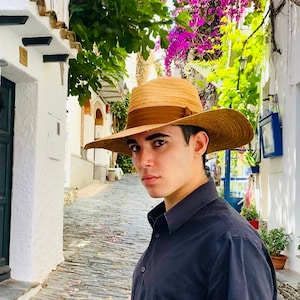 Peut inclure: Une jeune personne portant un chapeau de paille brun avec un ruban brun et une chemise boutonnée noire. La personne se tient dans une rue étroite pavée bordée de bâtiments blancs.