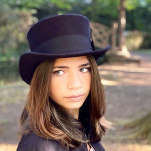 May include: A young person with long brown hair wearing a black top hat with a black band.