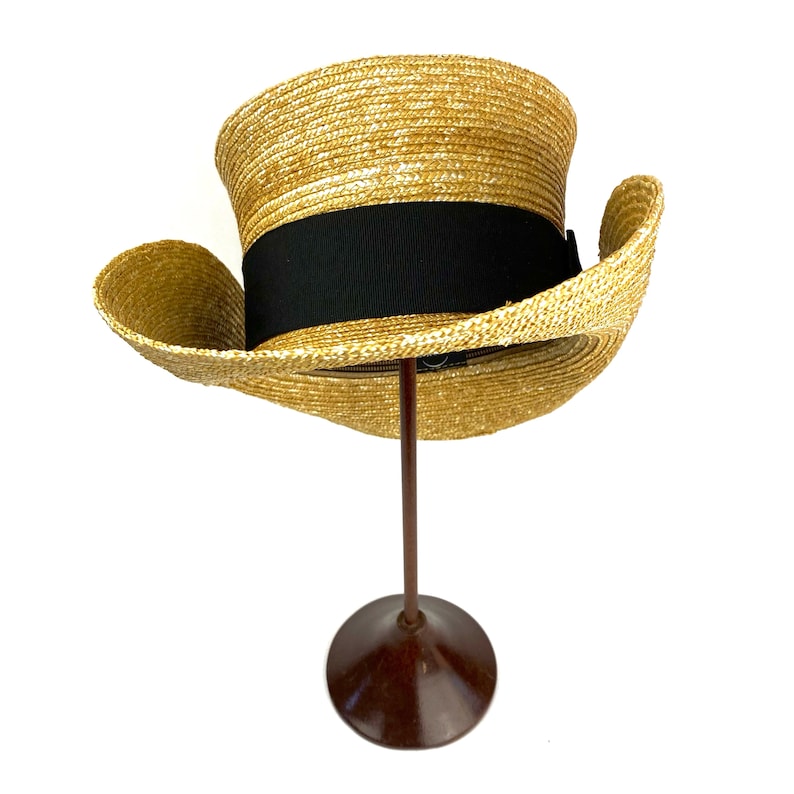 Pu&ograve; includere: Un cappello di paglia con un nastro nero. Il cappello &egrave; su un supporto marrone.