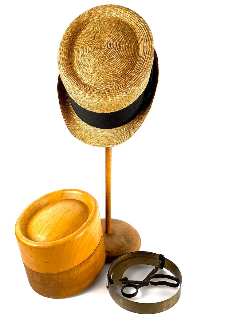 Peut inclure: Un chapeau de paille avec un ruban noir est pos&eacute; sur un support de chapeau en bois. &Agrave; c&ocirc;t&eacute; se trouve un bloc de chapeau en bois et un outil de mesure de chapeau en m&eacute;tal.