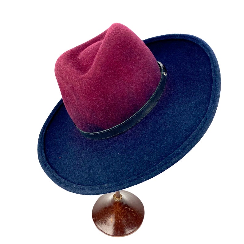 May include: A burgundy and navy blue fedora hat with a black leather band. The hat is sitting on a wooden hat stand.