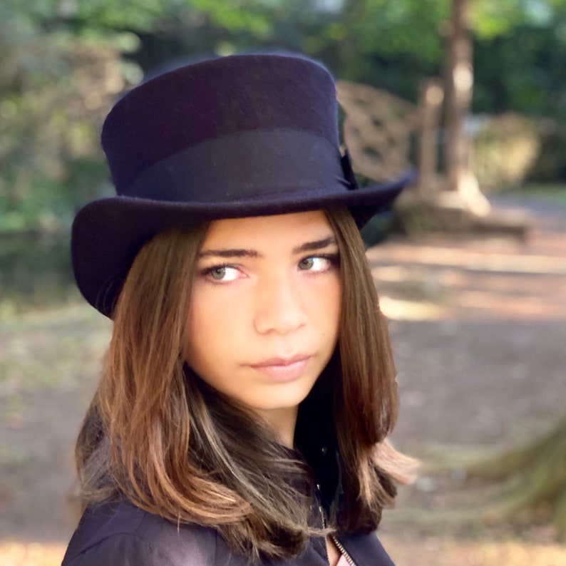 May include: A young person with long brown hair wearing a black top hat with a black band.