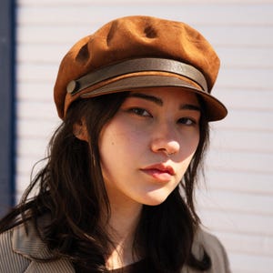 Puede incluir: Una gorra de estilo marinero en ante marrón con visera estructurada y una banda de aspecto de cuero marrón oscuro en la parte frontal. La banda está sujeta en el lateral con un botón circular, creando una silueta clásica y texturizada.