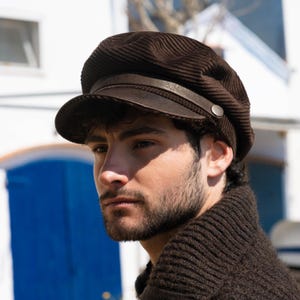 Puede incluir: Una gorra de estilo marinero en pana de color marrón oscuro con visera estructurada, una banda decorativa de aspecto cuero en la parte frontal y un pequeño botón metálico en el lateral. La gorra presenta una textura acanalada y copa suave.