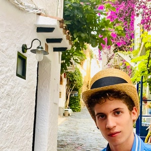 Peut inclure: Une jeune personne portant une chemise ray&eacute;e bleu et blanc et un chapeau de paille avec un bandeau noir. La personne se tient dans une rue &eacute;troite pav&eacute;e bord&eacute;e de b&acirc;timents blancs et de verdure luxuriante.