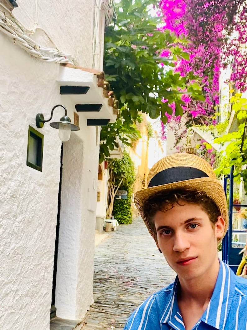 Peut inclure: Une jeune personne portant une chemise ray&eacute;e bleu et blanc et un chapeau de paille avec un bandeau noir. La personne se tient dans une rue &eacute;troite pav&eacute;e bord&eacute;e de b&acirc;timents blancs et de verdure luxuriante.