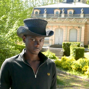 May include: A young man wearing a black top hat with a black and white striped band, a black mock neck shirt, and a gold embroidered logo on the left side of the shirt. He is standing in front of a large building with a white facade and a green lawn.