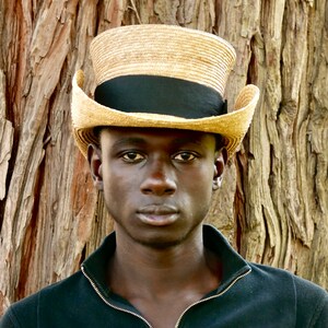 Pu&ograve; includere: Un uomo che indossa una camicia nera e un cappello di paglia a tesa larga con una fascia nera. Il cappello ha una corona alta e cilindrica.