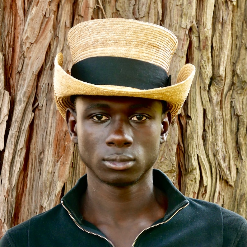Pu&ograve; includere: Un uomo che indossa una camicia nera e un cappello di paglia a tesa larga con una fascia nera. Il cappello ha una corona alta e cilindrica.