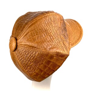 May include: A brown leather cap with a crocodile-embossed pattern. The cap has a curved brim and a button on the side.