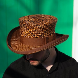 May include: A brown woven straw hat with a wide brim and a cylindrical crown. The hat has a dark brown band around the base of the crown. The hat is a classic style, suitable for sun protection or fashion.
