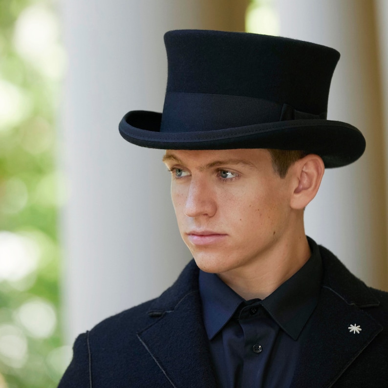 May include: A black top hat with a black band around the crown. The hat is worn by a person with a black jacket and a black shirt.