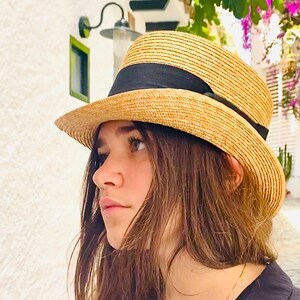 Peut inclure: Une femme aux longs cheveux bruns porte un chapeau de paille &agrave; larges bords avec un bandeau noir. Le chapeau a une couronne l&eacute;g&egrave;rement arrondie et une large bordure qui ombrage son visage. Elle regarde de c&ocirc;t&eacute; et son expression est pensive.