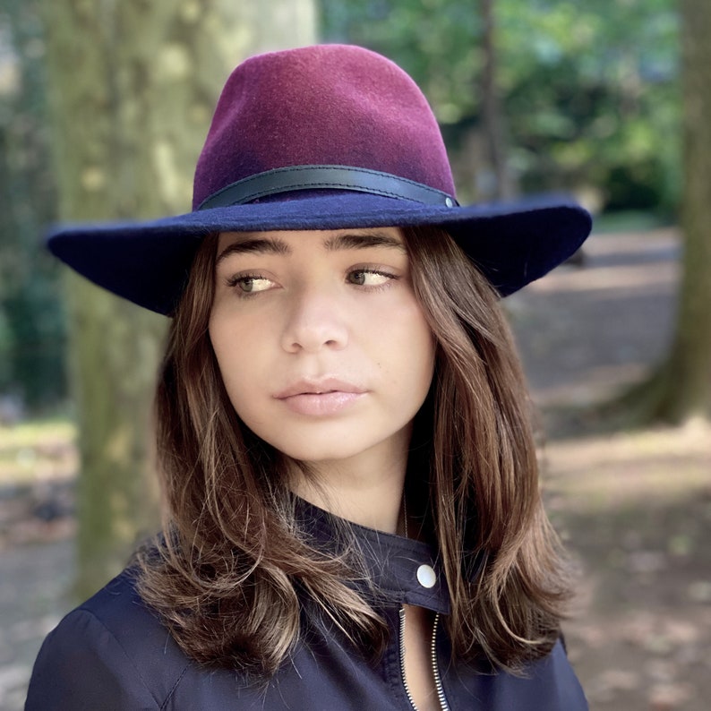 May include: A woman wearing a dark blue jacket with a zipper and a burgundy and navy blue fedora hat.