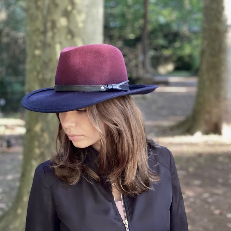 May include: A burgundy felt fedora with a black band and a black leather buckle. The hat has a wide brim and is worn by a person with long brown hair.
