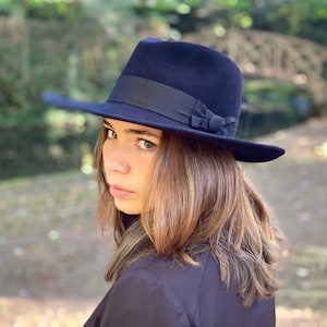 K&ouml;nnte beinhalten: Ein dunkelblauer Fedora-Hut mit einem schwarzen Band und einer kleinen Schleife. Der Hut wird von einer Person mit langen braunen Haaren getragen.