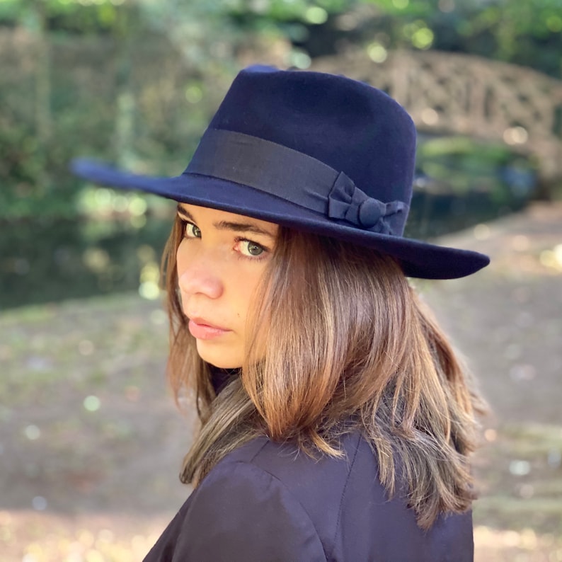 K&ouml;nnte beinhalten: Ein dunkelblauer Fedora-Hut mit einem schwarzen Band und einer kleinen Schleife. Der Hut wird von einer Person mit langen braunen Haaren getragen.