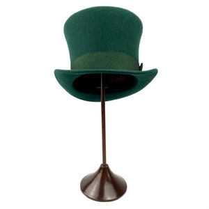 May include: A dark green top hat with a black band sits on a brown stand. The hat is made of felt and has a classic shape.