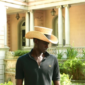 Pu&ograve; includere: Un uomo indossa una polo nera con un piccolo logo sul petto sinistro. Indossa un cappello di paglia a tesa larga con una fascia nera.