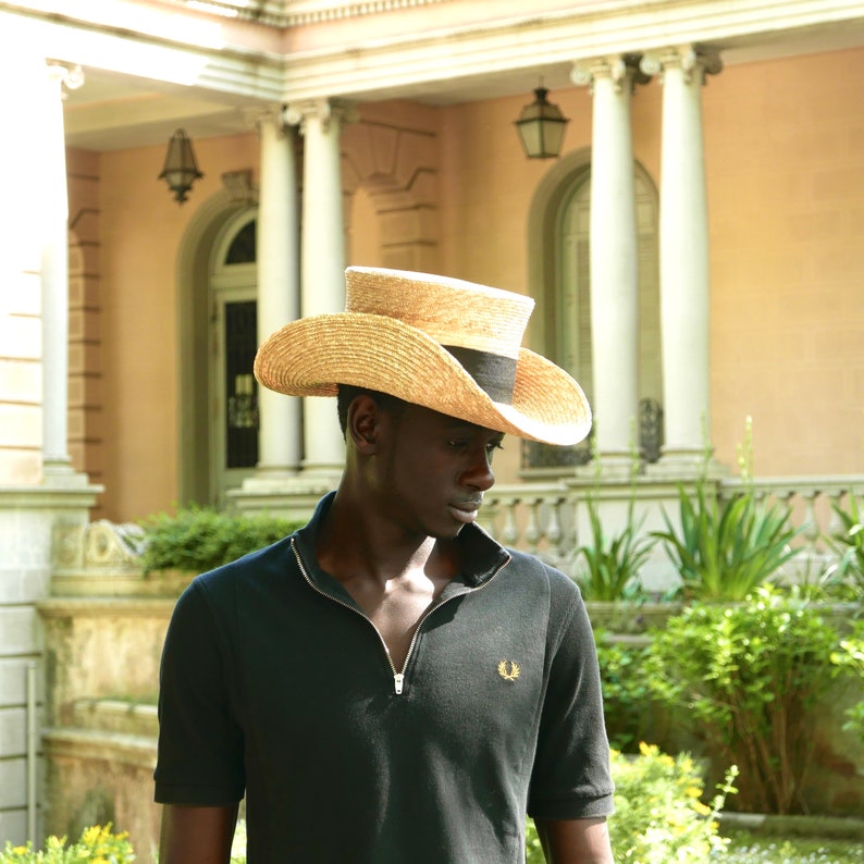 Pu&ograve; includere: Un uomo indossa una polo nera con un piccolo logo sul petto sinistro. Indossa un cappello di paglia a tesa larga con una fascia nera.
