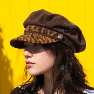May include: A brown newsboy cap featuring a leopard print brim and band. The cap has a round crown and a small, stiff brim. A gold-coloured button is on the side. The background is bright yellow.