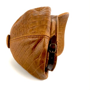 May include: A brown leather cap with a crocodile-embossed pattern and a brown leather strap with a brass buckle.