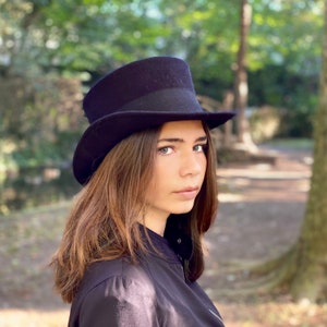 May include: A woman wearing a black top hat and a black jacket. She has long brown hair and is looking to the side.