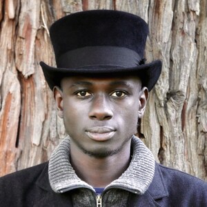 Peut inclure: Un homme portant un chapeau haut de forme noir et un pull gris sur une veste noire. Il se tient devant un tronc d'arbre.