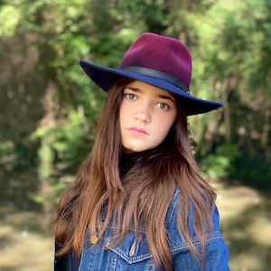 May include: A young person with long brown hair wearing a dark blue denim jacket and a burgundy fedora with a black band.