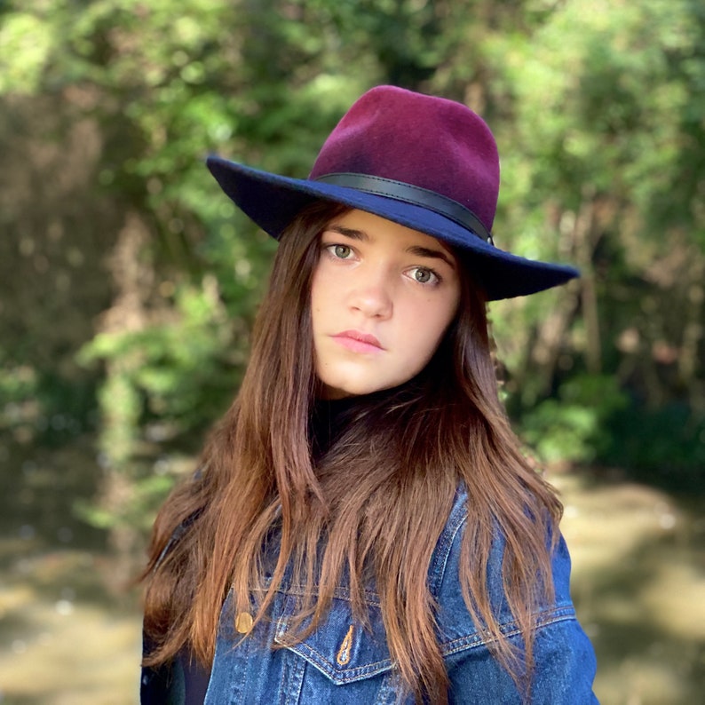 May include: A young person with long brown hair wearing a dark blue denim jacket and a burgundy fedora with a black band.