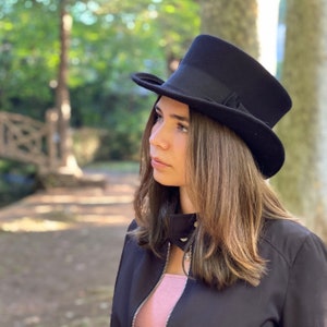 May include: A woman wearing a black top hat and a black jacket. She has long brown hair and is looking off to the side.