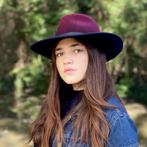 May include: A woman with long brown hair wearing a dark blue denim jacket and a burgundy fedora with a black band.