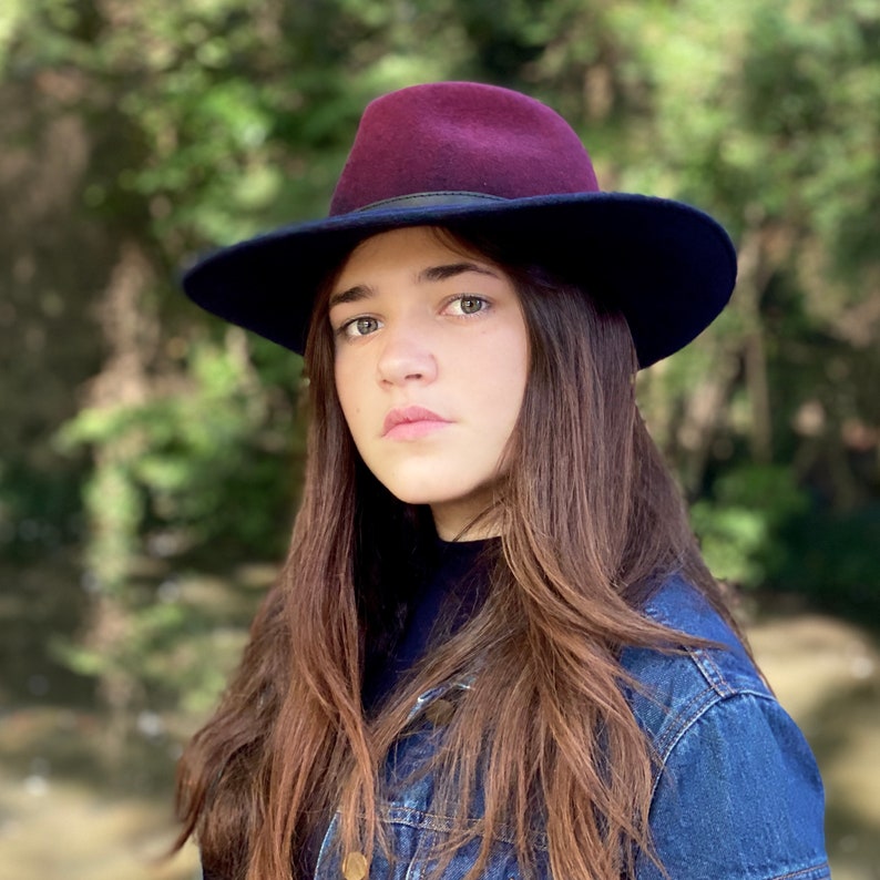 May include: A woman with long brown hair wearing a dark blue denim jacket and a burgundy fedora with a black band.