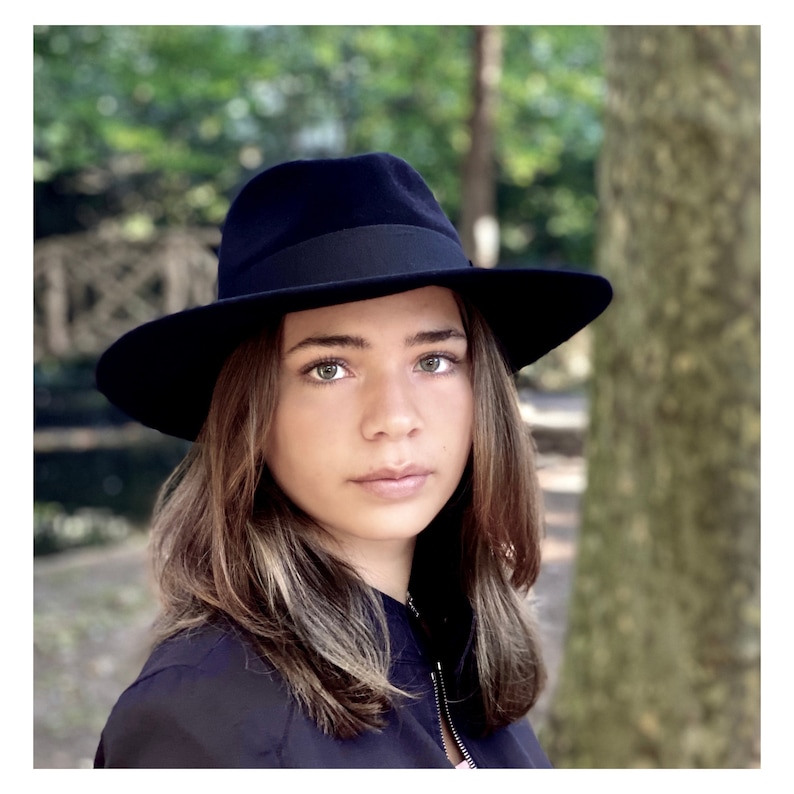 K&ouml;nnte beinhalten: Eine junge Person mit langen braunen Haaren, die einen schwarzen Fedora-Hut und eine schwarze Jacke tr&auml;gt.