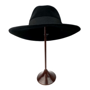 K&ouml;nnte beinhalten: Ein schwarzer Fedora-Hut mit breiter Krempe, der auf einem braunen St&auml;nder steht.