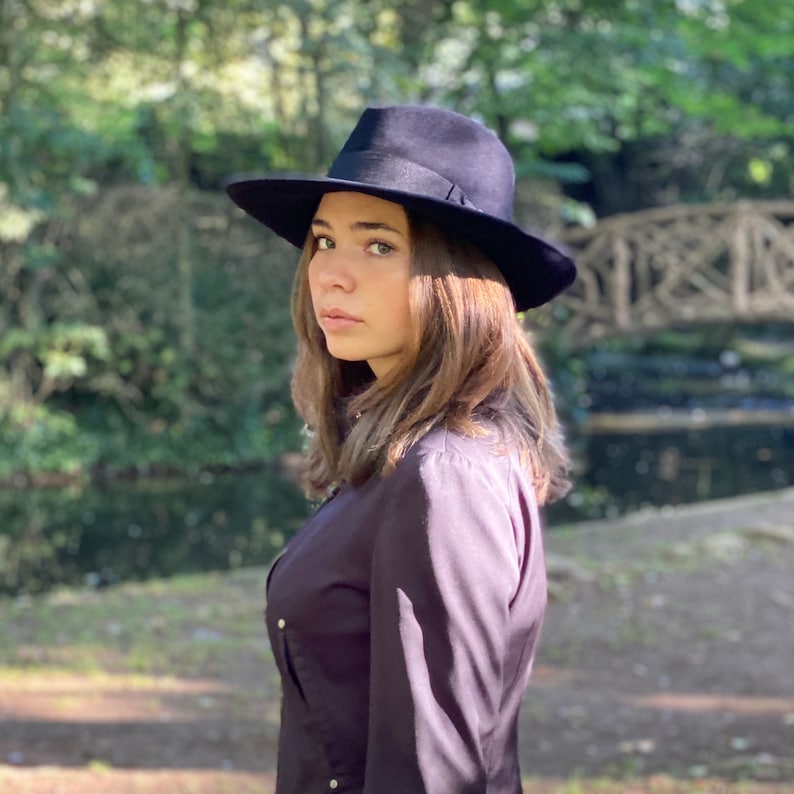 K&ouml;nnte beinhalten: Eine Frau tr&auml;gt einen schwarzen Fedora-Hut und ein dunkelviolettes Langarmhemd. Sie steht in einem Park mit einer Holzbr&uuml;cke im Hintergrund.