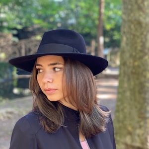 K&ouml;nnte beinhalten: Eine Frau tr&auml;gt einen schwarzen Fedora-Hut mit einem schwarzen Band. Sie tr&auml;gt eine schwarze Jacke und hat lange braune Haare.