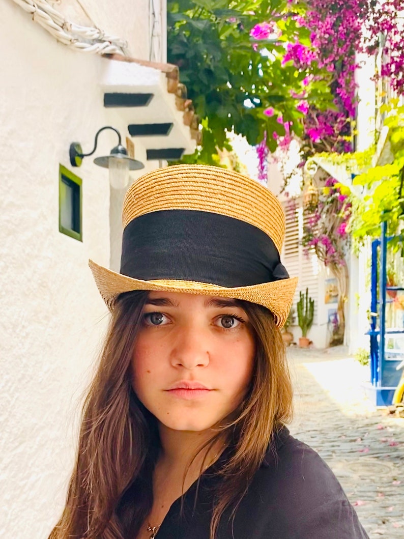Peut inclure: Une femme aux longs cheveux bruns porte un chapeau de paille avec un bandeau noir. Le chapeau a une couronne haute et cylindrique et une large bordure. Elle regarde l'appareil photo avec une expression s&eacute;rieuse.