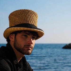 May include: A straw top hat with a black band. The hat has a woven texture and a tall crown. The person is wearing a black shirt. The background is a blue ocean and sky.