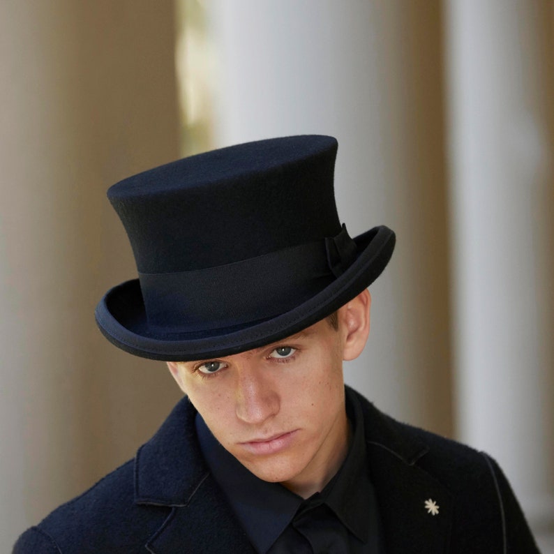 May include: A black top hat with a wide brim and a black band around the crown. The hat is worn by a person with a black jacket and a black shirt.