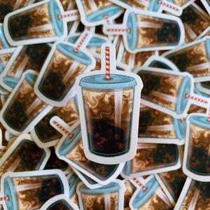 May include: A close-up of a stack of stickers featuring a cartoon illustration of a iced coffee drink with a red and white striped straw. The stickers are printed on white paper and have a glossy finish.