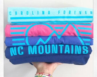 May include: Stack of four t-shirts with different designs. The top shirt is light blue with the text "CAROLINA FOREVER" in turquoise. The second shirt is pink with a turquoise mountain design. The third shirt is purple with a turquoise mountain design. The bottom shirt is navy blue with the text "NC MOUNTAINS" in turquoise.
