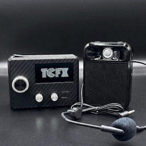 May include: Black audio equipment set with a carbon fiber texture. The set includes a rectangular device with a screen displaying "TCFX", a control knob, and two buttons. A second, smaller device has a microphone and a headset.