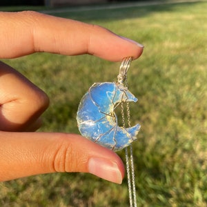 May include: A silver necklace featuring a crescent moon pendant. The moon is crafted from a light blue, translucent stone, wrapped in silver wire. The necklace hangs on a delicate silver chain, creating a celestial-themed accessory.