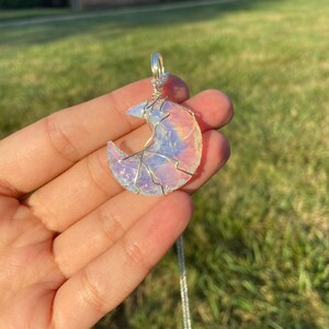 May include: A crescent moon pendant with a silver chain. The moon is made of iridescent, opalescent material, wrapped in silver wire. The pendant is held in a hand against a backdrop of green grass and sunlight.