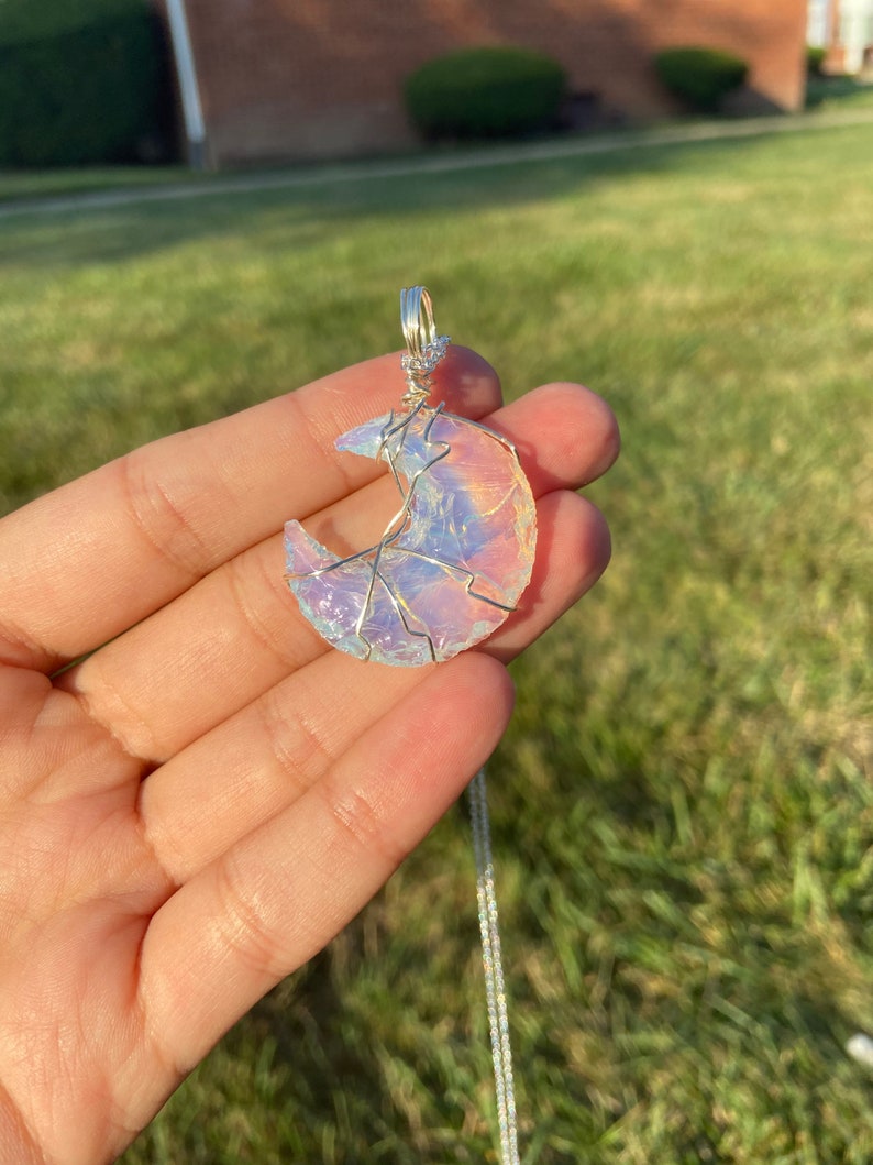 May include: A crescent moon pendant with a silver chain. The moon is made of iridescent, opalescent material, wrapped in silver wire. The pendant is held in a hand against a backdrop of green grass and sunlight.