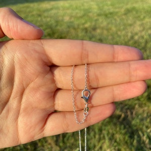 May include: A delicate silver necklace with a small clasp and chain, held in a hand against a blurred green background. The necklace is simple and elegant, perfect for everyday wear. The chain is thin and the clasp is small.