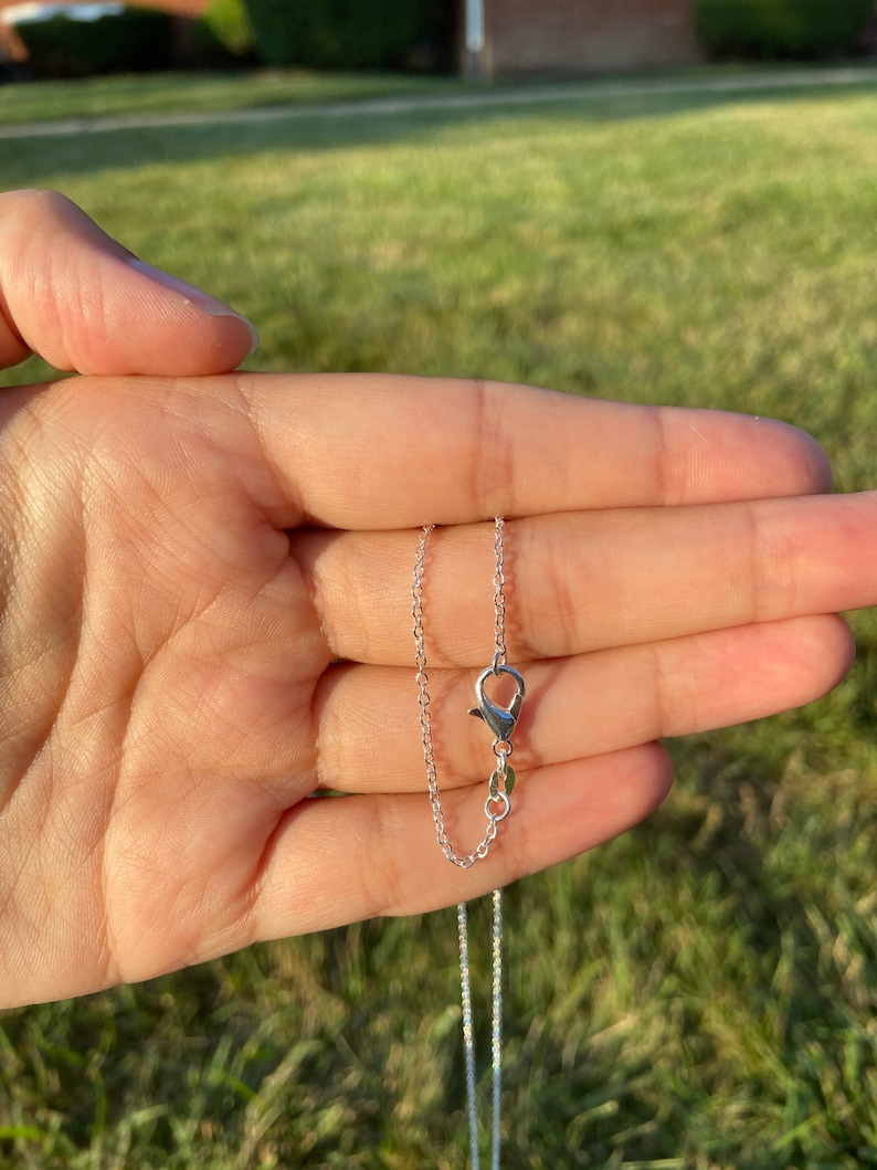 May include: A delicate silver necklace with a small clasp and chain, held in a hand against a blurred green background. The necklace is simple and elegant, perfect for everyday wear. The chain is thin and the clasp is small.