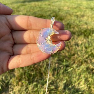 May include: A crescent moon pendant necklace. The moon-shaped pendant is made of a translucent, iridescent material, wrapped in silver-colored wire. The necklace chain is also silver-colored. The pendant is held up by a hand against a background of green grass.